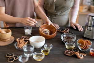 Young woman mixing ground candied fruits in wooden bowl while standing by table with ingredients for making natural soap