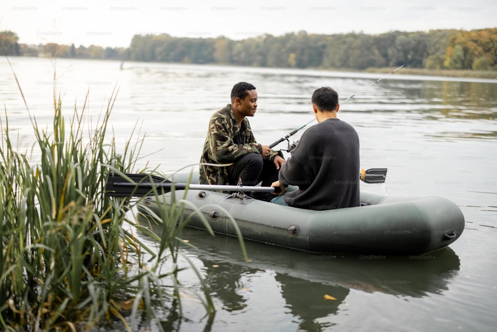 Multiracial male friends fishing with fishing rods on rubber boat in lake or river. Concept of rest, weekend and hobby in nature. Idea of friendship and spending time together. Autumn day