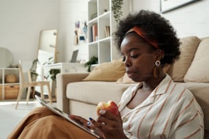 Vielbeschäftigter junger afrikanischer Freiberufler mit Apfel, der im Tablet scrollt, während er zu Hause im Wohnzimmer netzwerkt