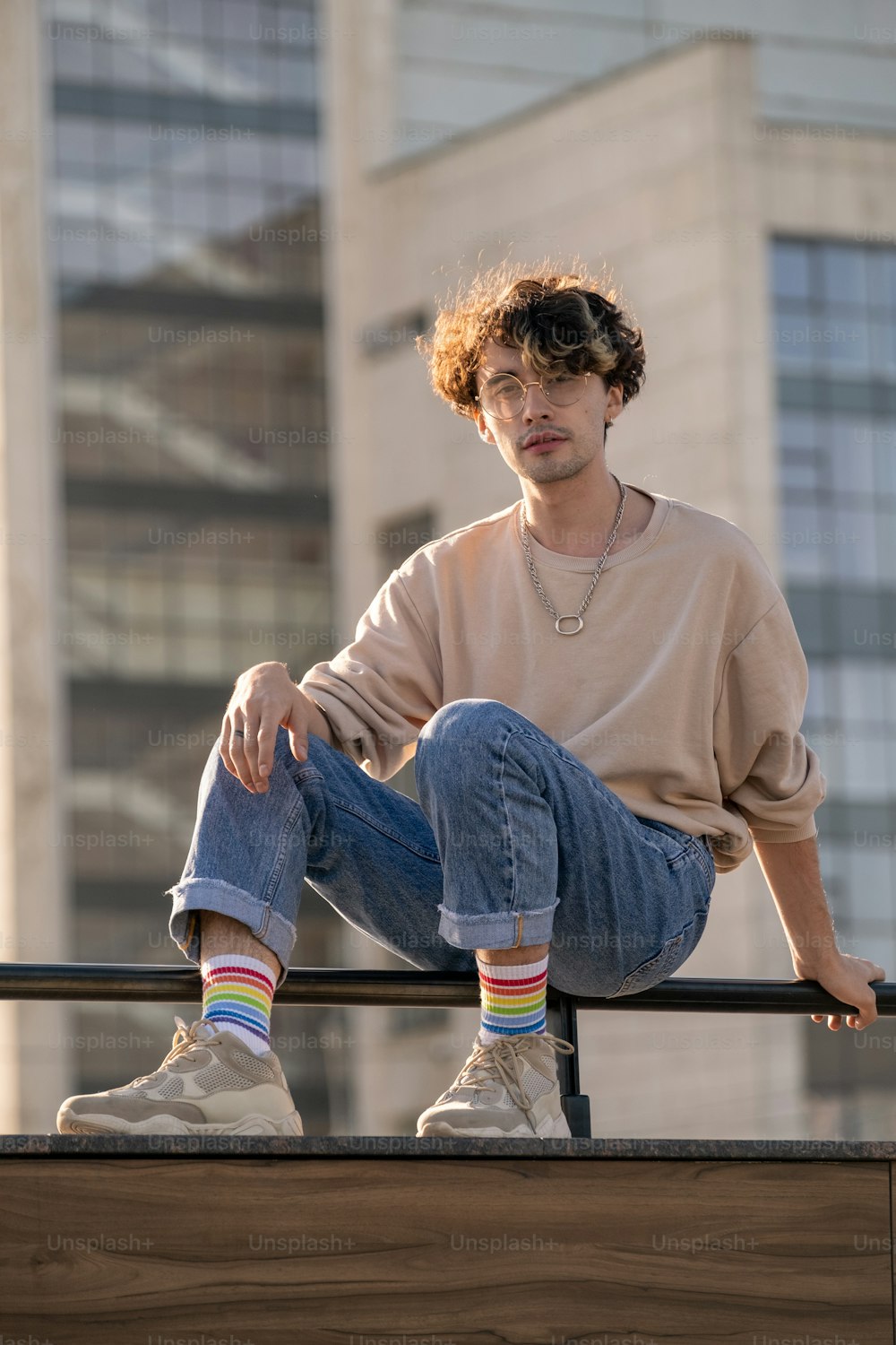 Contemporary teenage guy in jeans and sweatshirt sitting on railings against building exterior