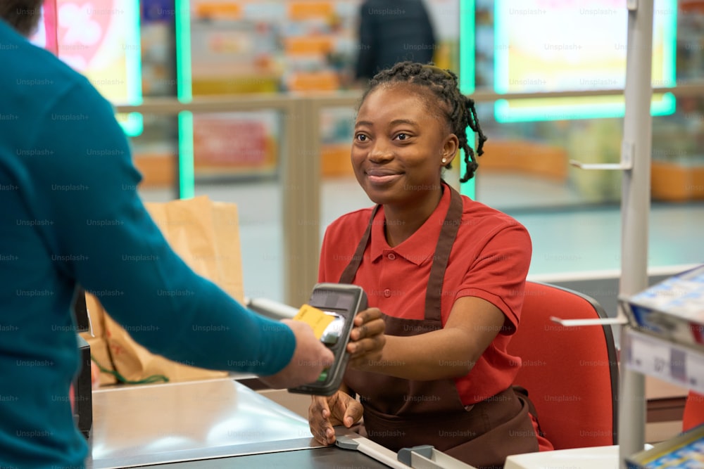 スーパーマーケットのレジカウンターのそばに座って支払い端末を保持しながら、クレジットカードで支払う購入者を見ている幸せな販売員