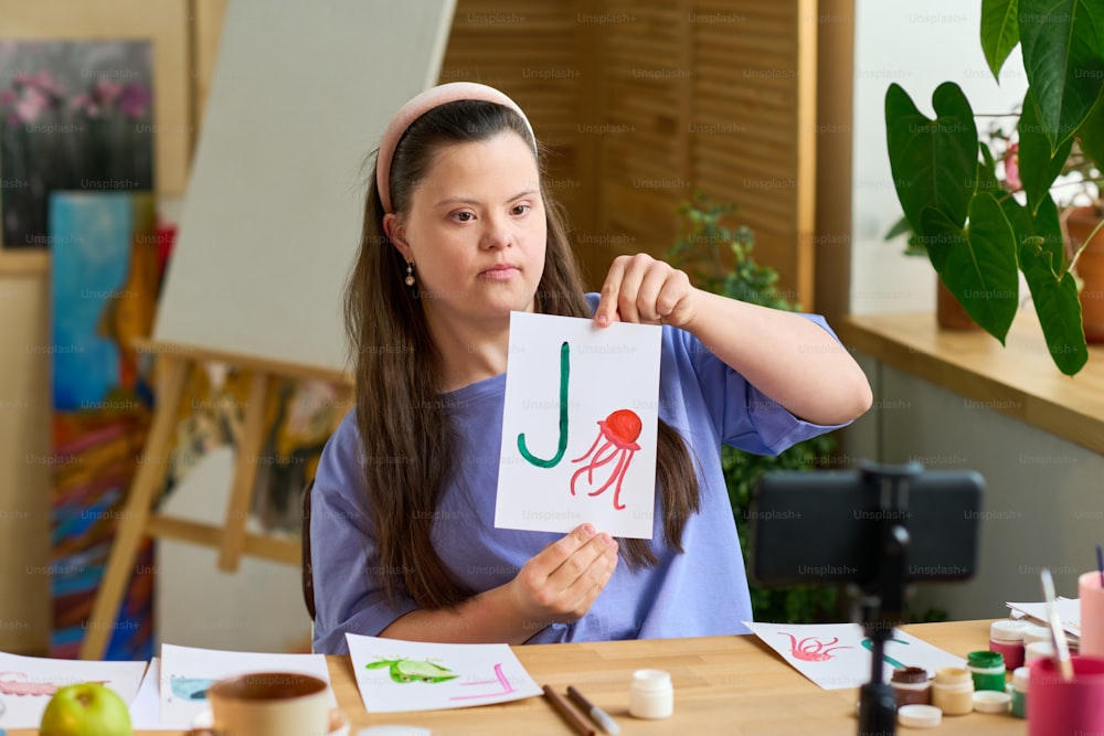 Menina com síndrome de Down mostrando papel com letra inglesa e desenho de águas-vivas para o público on-line enquanto está sentada na frente da câmera