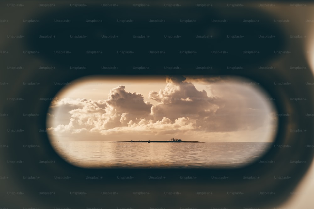 View through opened vessel porthole of a tiny island surrounded by ocean water in the evening, with silhouettes of people on it and a stunning sunset cloudscape above, Maldives