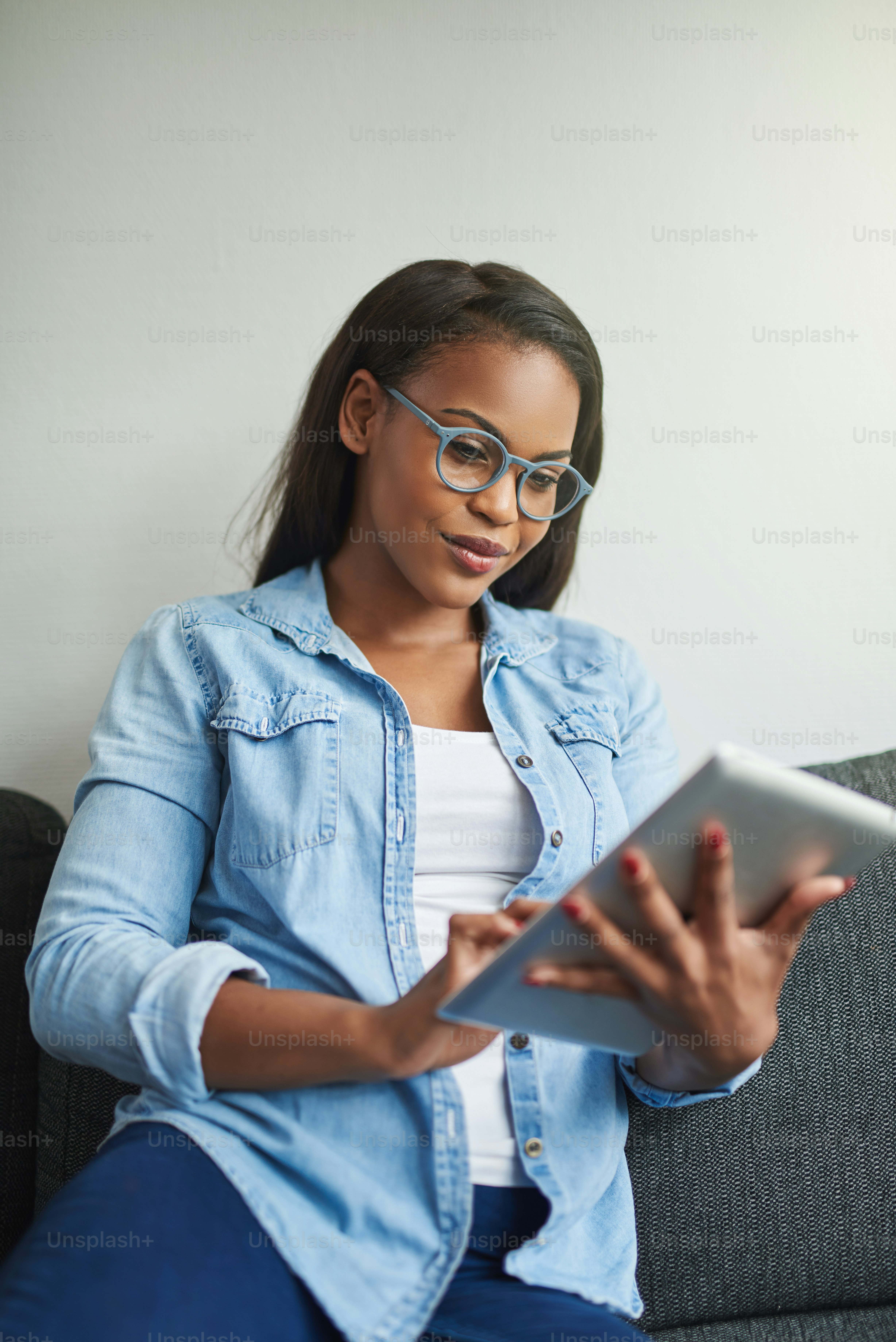 Young African woman wearing glasses relaxing on the sofa in her living room browsing online with a digital tablet