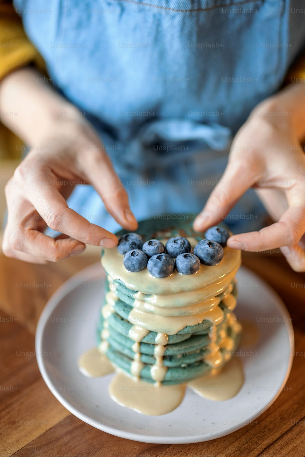 青いエプロンを着た女性は、木製のテーブルにキャラメルのスーズを注いだブルーベリーで青いアメリカのパンケーキを飾ります。自家製のおいしい料理。シュローブタイドの祭典。