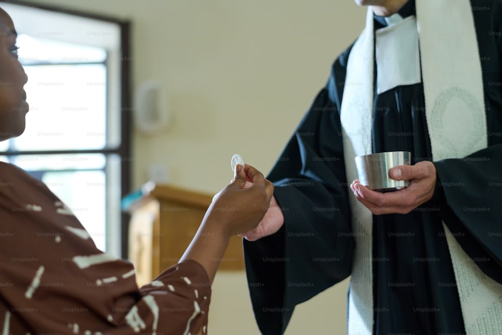 Young African American parishioner taking unleavened bread symbolizing flesh of Lord Jesus from hand of priest of Catholic church