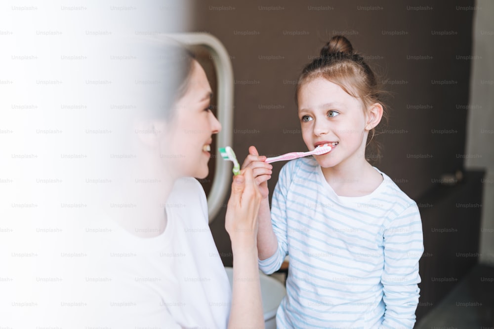 Young mother woman with long hair with little tween girl daughter in pajamas brushing their teeth in the morning at home