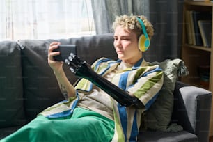 Junge ernste Frau mit myoelektrischem Arm, die das Smartphone vor sich hält, während sie im Video-Chat auf einem bequemen Sofa kommuniziert
