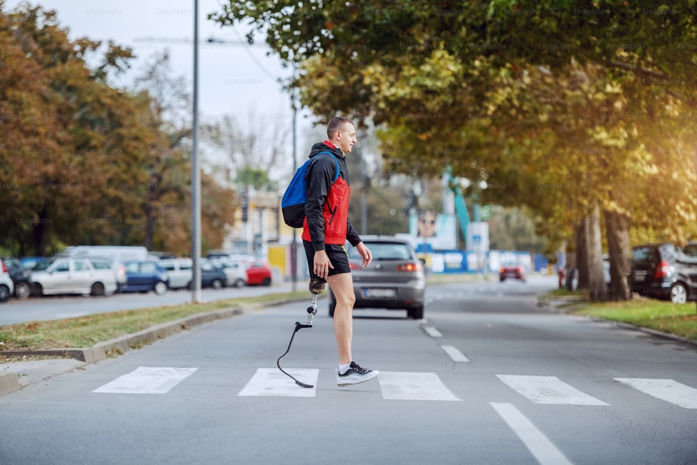 De cuerpo entero de guapo deportista caucásico discapacitado en ropa deportiva, con pierna artificial y mochila cruzando la calle.
