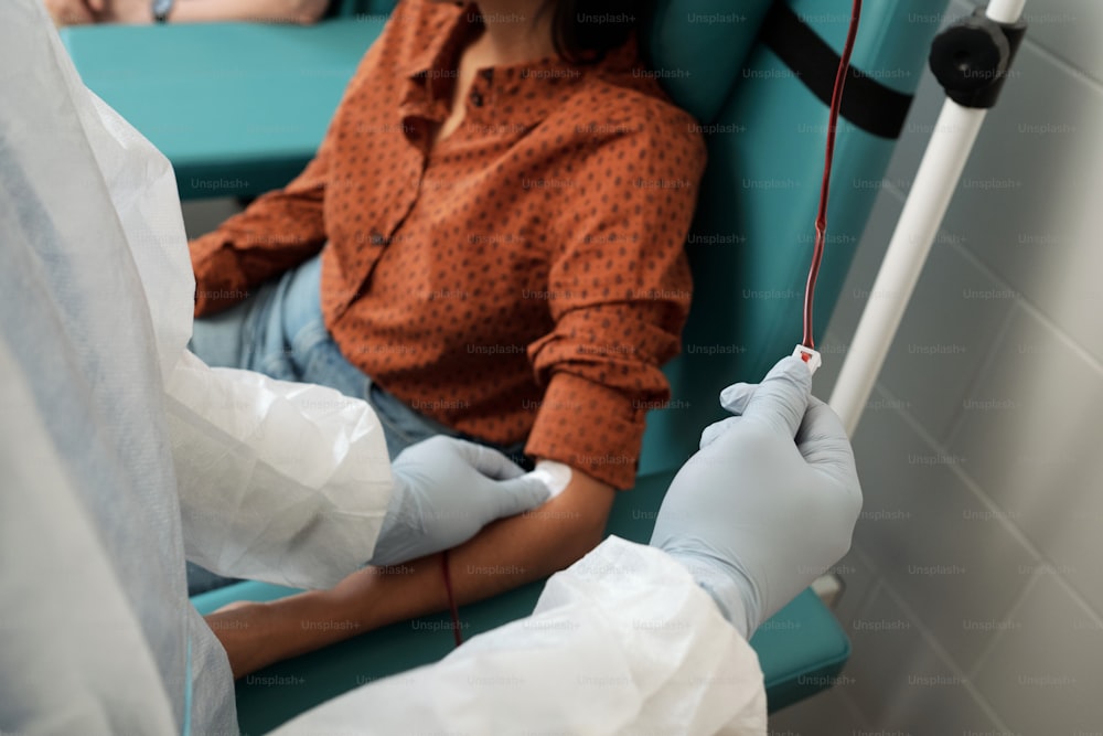 Behandschuhte Krankenschwester in Overalls setzt Tropferschlauch in die Vene einer Spenderin oder Freiwilligen, die während des Hämotransfusionsverfahrens auf dem Stuhl sitzt
