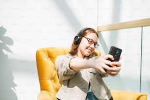 携帯電話を使用してメガネで若いブルネットのティーンエイジャーの女の子の大学生または高校生は、公共の場で黄色い椅子でヘッドフォンで音楽を聴く