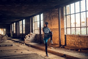 Portrait d’un jeune homme athlétique afro-américain actif et attrayant avec des écouteurs, debout et faisant de l’entraînement d’étirement avec la jambe levée à l’intérieur de l’endroit abandonné.