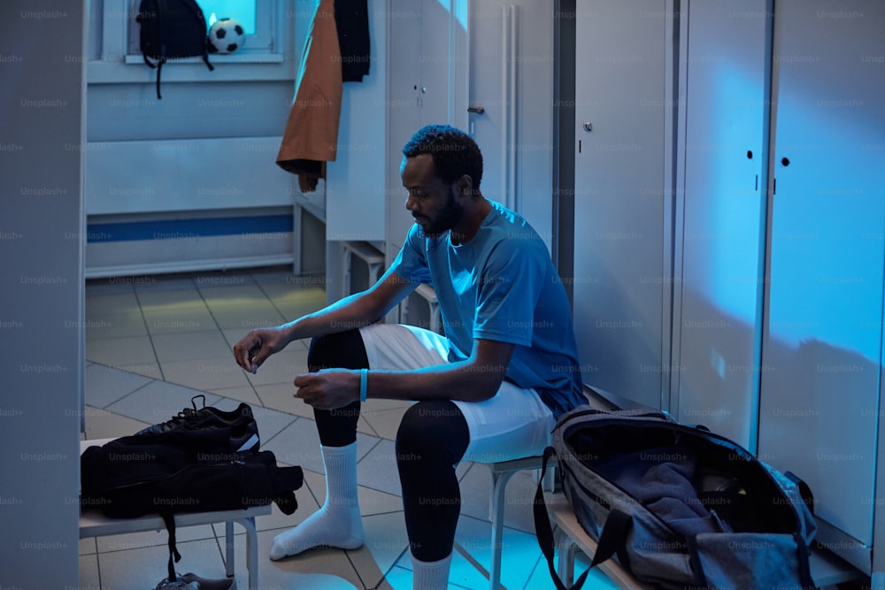 Jeune footballeur africain qui va enfiler ses chaussures de sport assis près d’une rangée de casiers dans les vestiaires