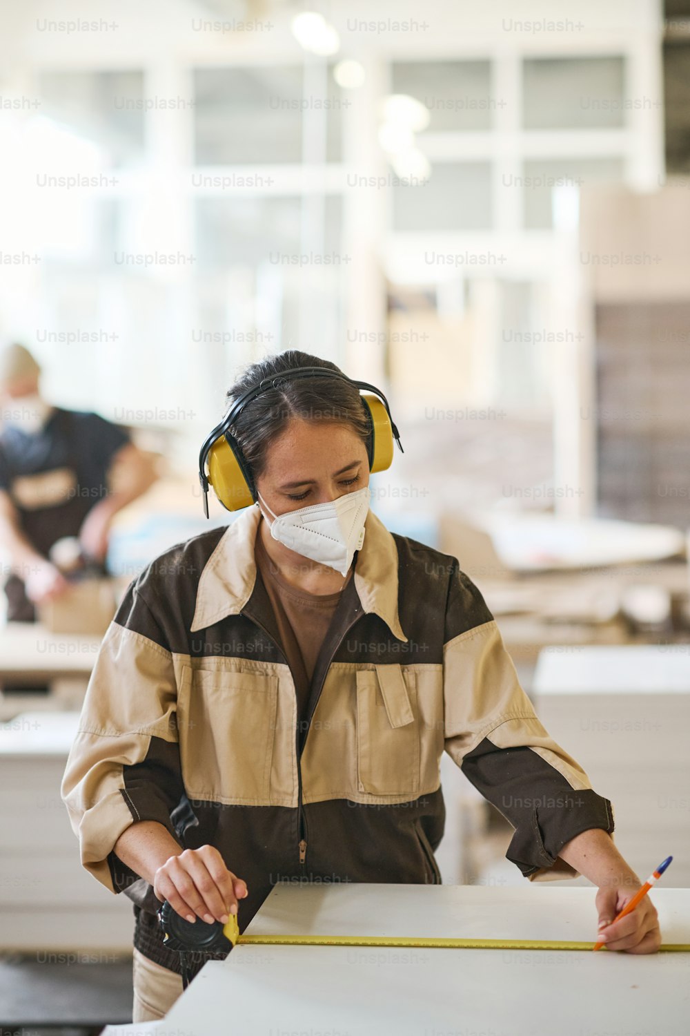 Female carpenter in protective mask and headphones making measurements of wooden boards with ruler at factory