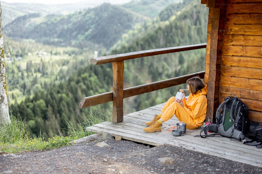 A jovem desfruta de uma excelente paisagem montanhosa e come comida sublimada para caminhadas, enquanto se senta relaxada em um terraço de madeira. Conceito de comida para viagens e fugas para a natureza