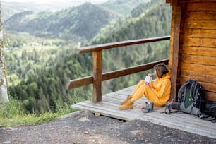 Junge Frau genießt die tolle Berglandschaft und isst sublimiertes Essen zum Wandern, während sie entspannt auf einer Holzterrasse sitzt. Konzept der Nahrung für Reisen und Flucht in die Natur