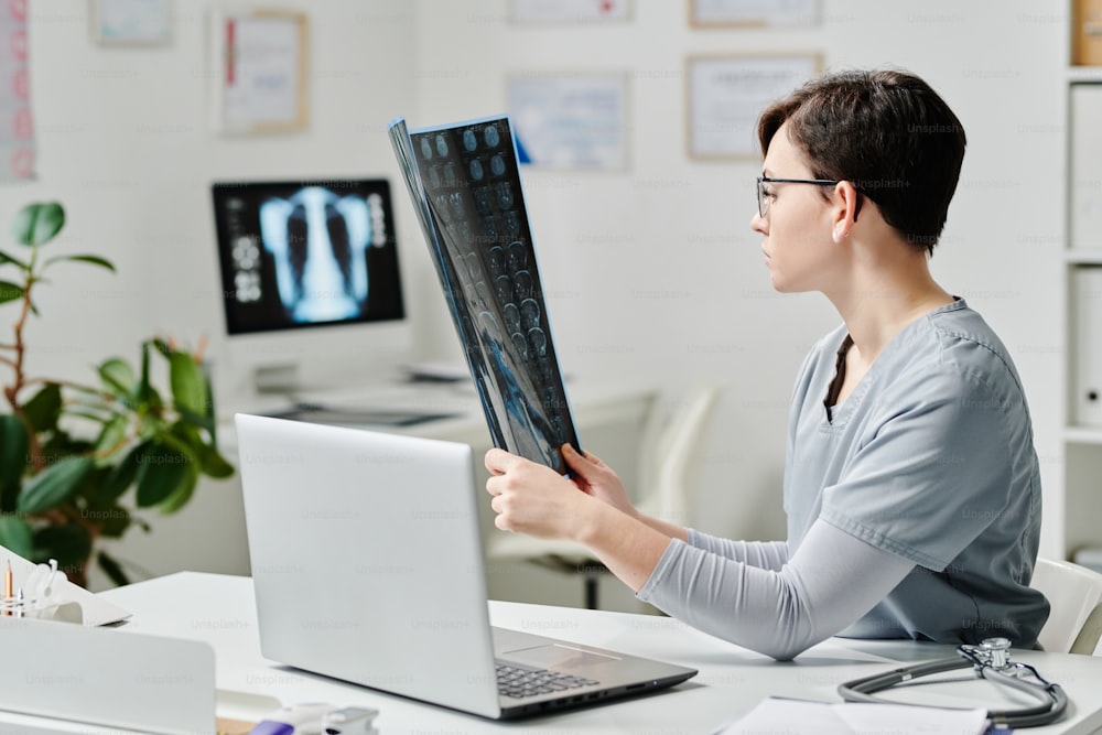 Young serious female clinicia in blue medical scrubs learning x-ray image of patient during online consultation by workplace in office