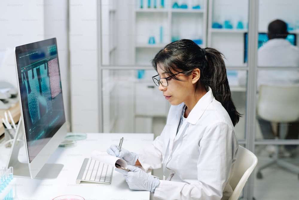 Contemporary young female researcher in whitecoat and gloves making scientific notes in notepad while sitting in front of computer