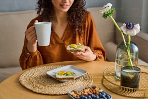 Brünettes Mädchen mit Tasse Kräutertee und leckerem vegetarischem Sandwich, das am servierten Küchentisch sitzt und das selbstgemachte Frühstück genießt