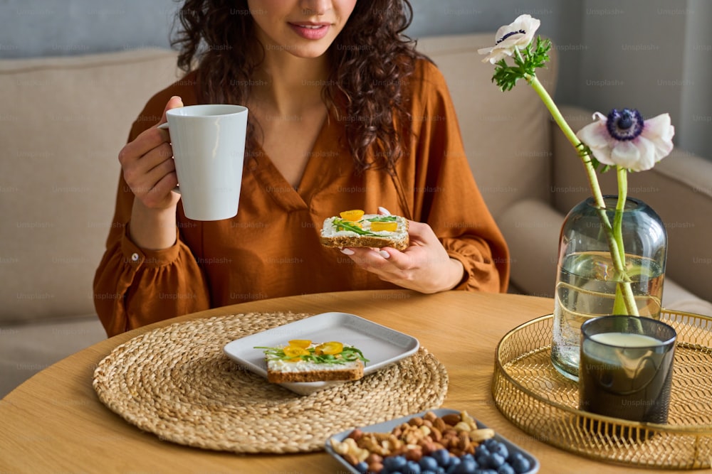 허브 차 한잔과 맛있는 채식 샌드위치를 가진 갈색 머리 소녀는 식탁에 앉아 스스로 만든 아침 식사를 즐기고 있습니다.