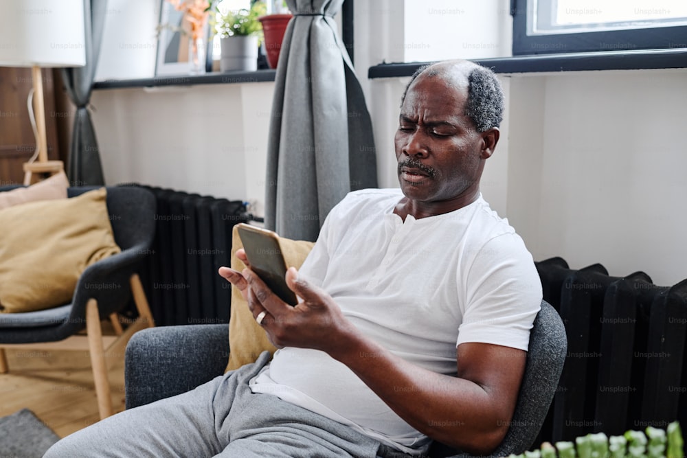Homem negro aposentado sério olhando para a tela do smartphone durante a comunicação por mensagens ou em bate-papo por vídeo em casa