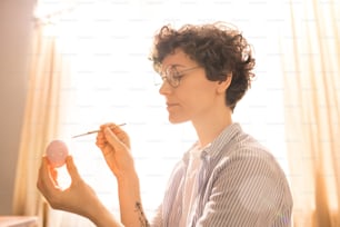 Giovane donna casuale in occhiali da vista che fa punti con vernice bianca su uovo di Pasqua rosa chiaro
