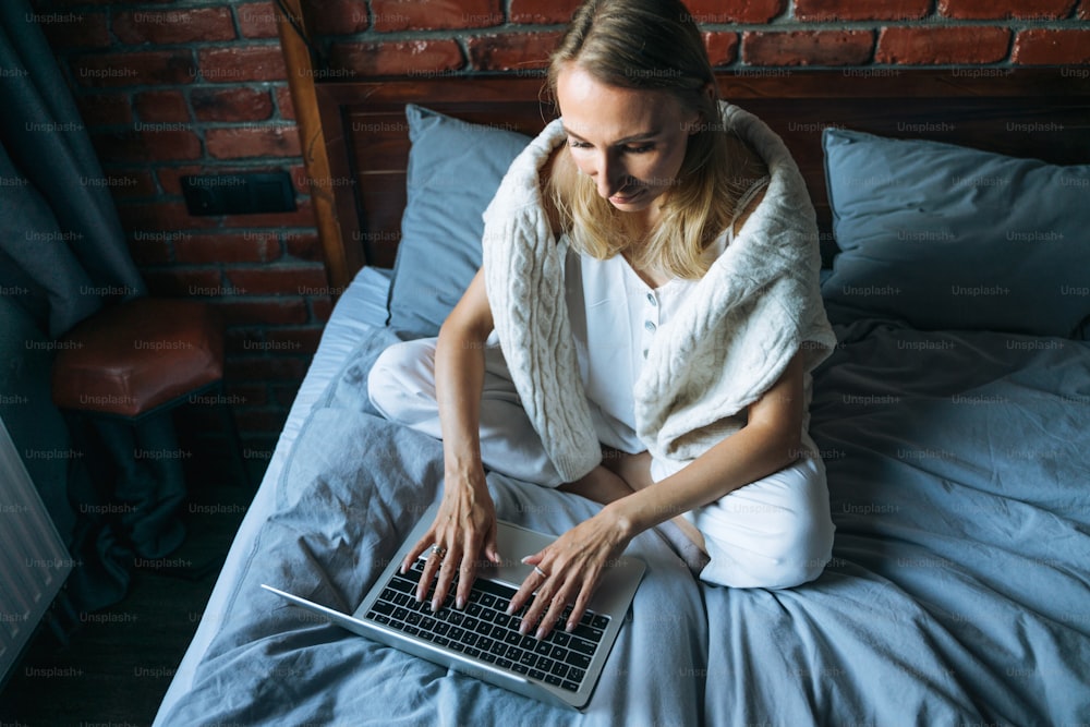 Mujer adulta de cuarenta años freelancer con cabello largo rubio en ropa informal usando computadora portátil sentada en la cama en casa, vista desde arriba