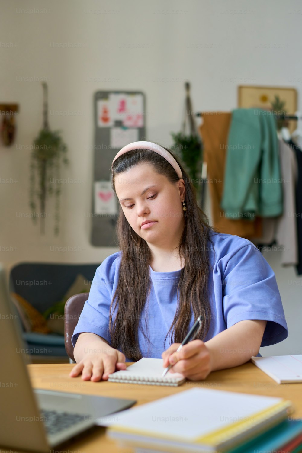 Female student with Down syndrome making notes in notepad during online lesson while sitting by desk in front of laptop at home