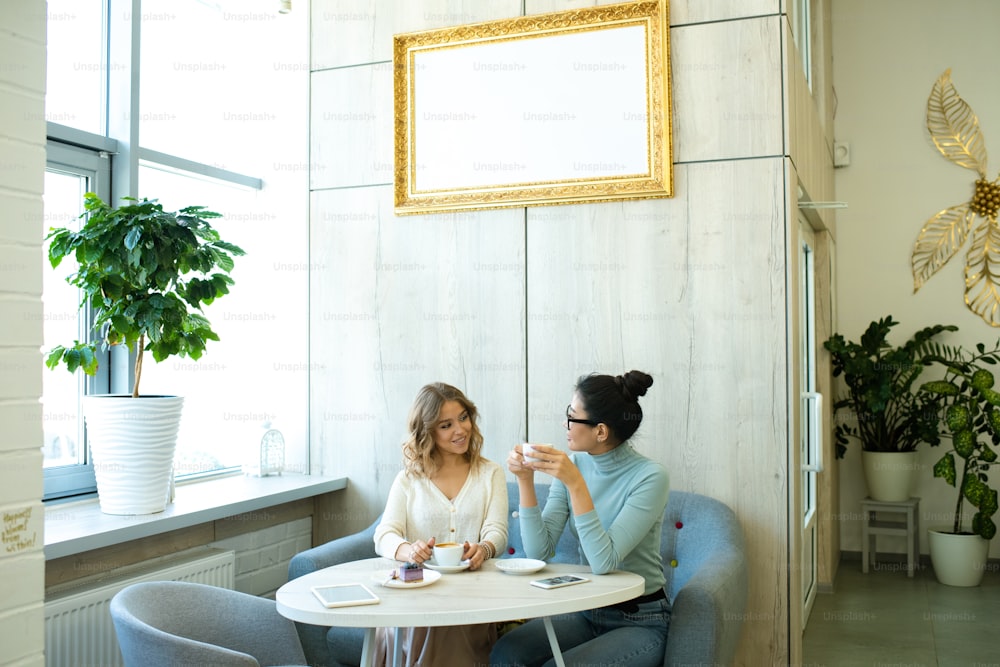Dos mujeres jóvenes amigables en ropa informal sentadas en un sofá suave y cómodo junto a la mesa en la cafetería, tomando café e interactuando