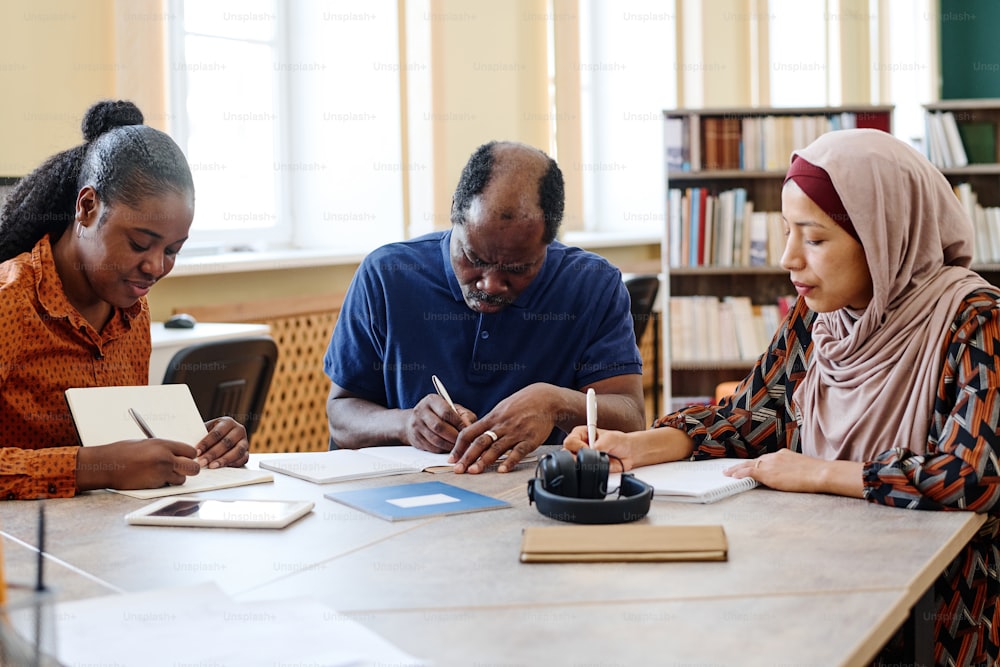 Grupo de tres estudiantes inmigrantes multiétnicos sentados a la mesa en la biblioteca haciendo tareas de escritura durante la lección