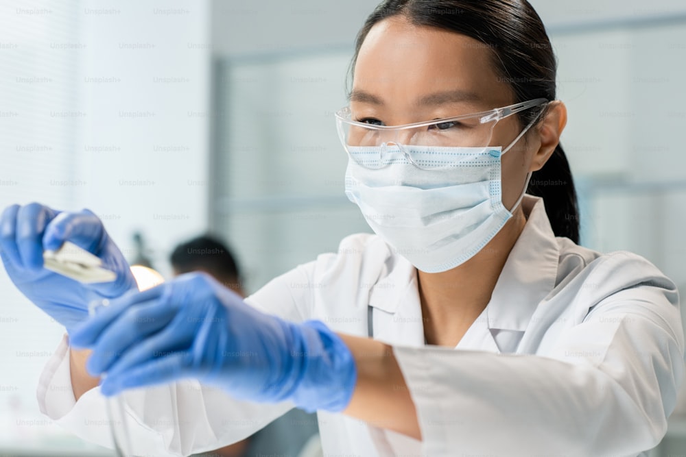 白衣とマスクを着用し、手袋をはめた若い女性研究者が、実験室での実験中に食品成分の小さなサンプルをフラスコに入れる