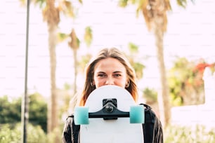 Modern cheerful beautiful blonde girl portrait with skateboard covering part of the pretty face - young millennial people outdoor concept enjoying lifestyle and sunny days