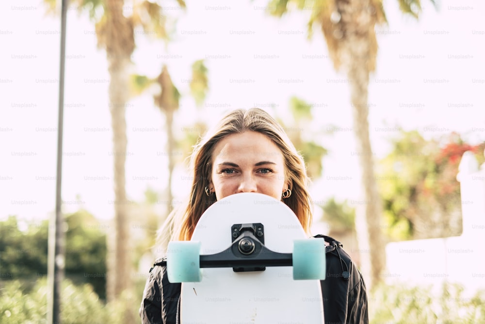 Modern cheerful beautiful blonde girl portrait with skateboard covering part of the pretty face - young millennial people outdoor concept enjoying lifestyle and sunny days