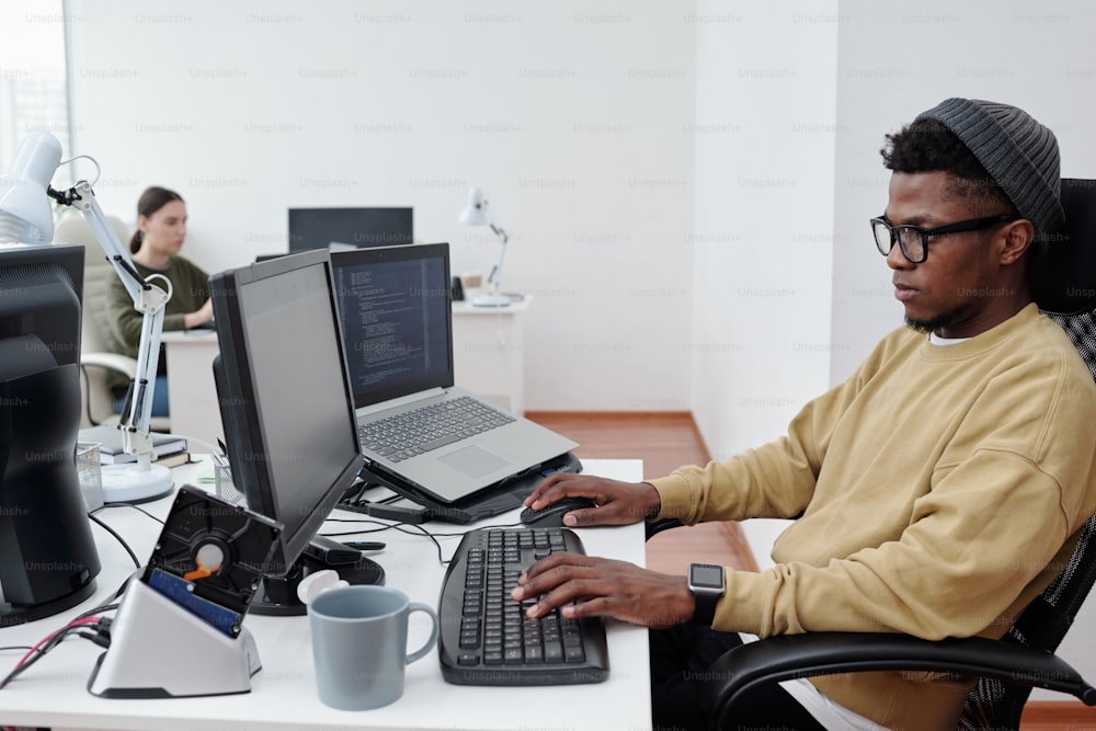 Jeune développeur informatique afro-américain en tenue décontractée décodant des données assis devant un écran d’ordinateur dans un bureau moderne en openspace