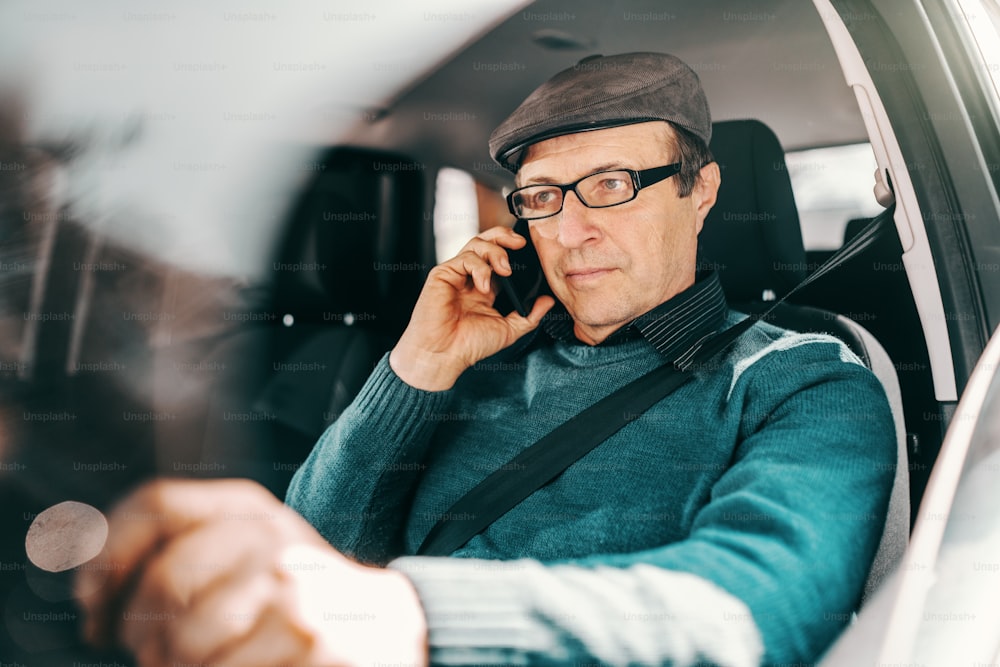 Serious Caucasian senior man with cap on head and eyeglasses sitting in car with hand on steering wheel and using smart phone. Window opened.