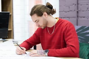 Young serious designer sitting at table and drawing construction project with pencil