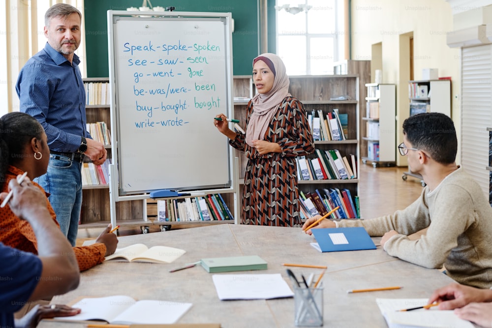 Muslim woman wearing hijab standing at whiteboard doing task with irregular verbs during English lesson