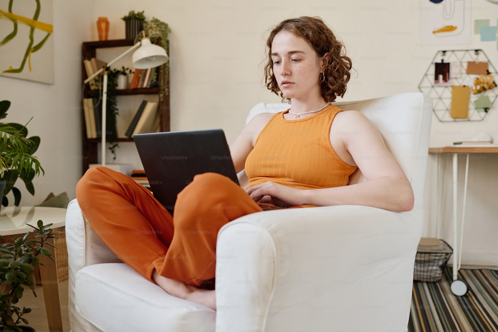 Joven empresaria seria con una laptop de rodillas concentrándose en la red mientras se sienta en un cómodo sillón de cuero blanco