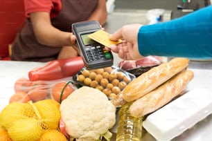 Hand eines männlichen Verbrauchers, der die Kreditkarte über den Bildschirm des Zahlungsterminals hält, das von einer afroamerikanischen Verkäuferin über Lebensmittelprodukten gehalten wird