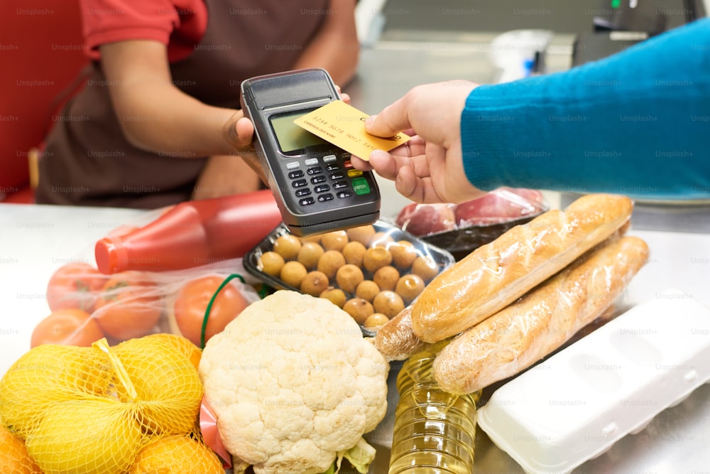 Mano del consumatore maschio che tiene la carta di credito sopra lo schermo del terminale di pagamento tenuto dalla commessa afroamericana sui prodotti alimentari