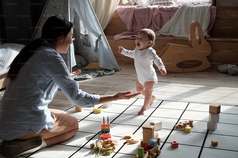 Jovem mãe amorosa vestindo roupas casuais sentada no chão da sala de estar incentivando seu bebê a caminhar em direção a ela