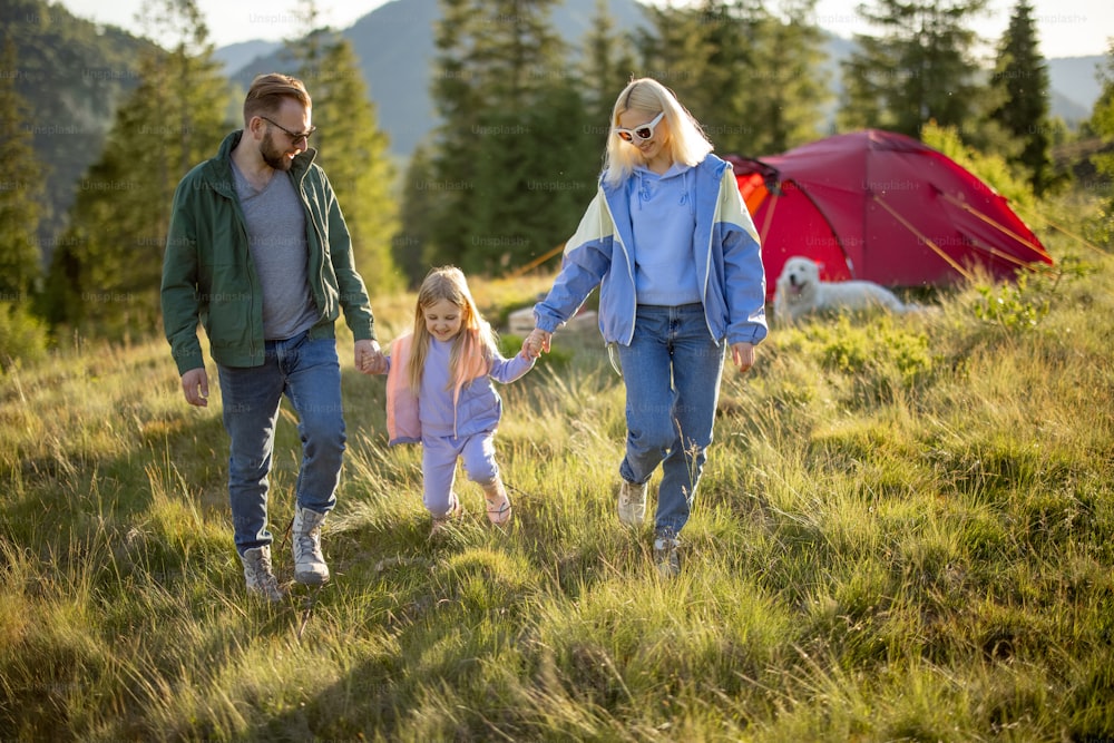 Young caucasian family with little girl walk together on green meadow while traveling with tent in the mountains. Happy family summer vacation at campsite