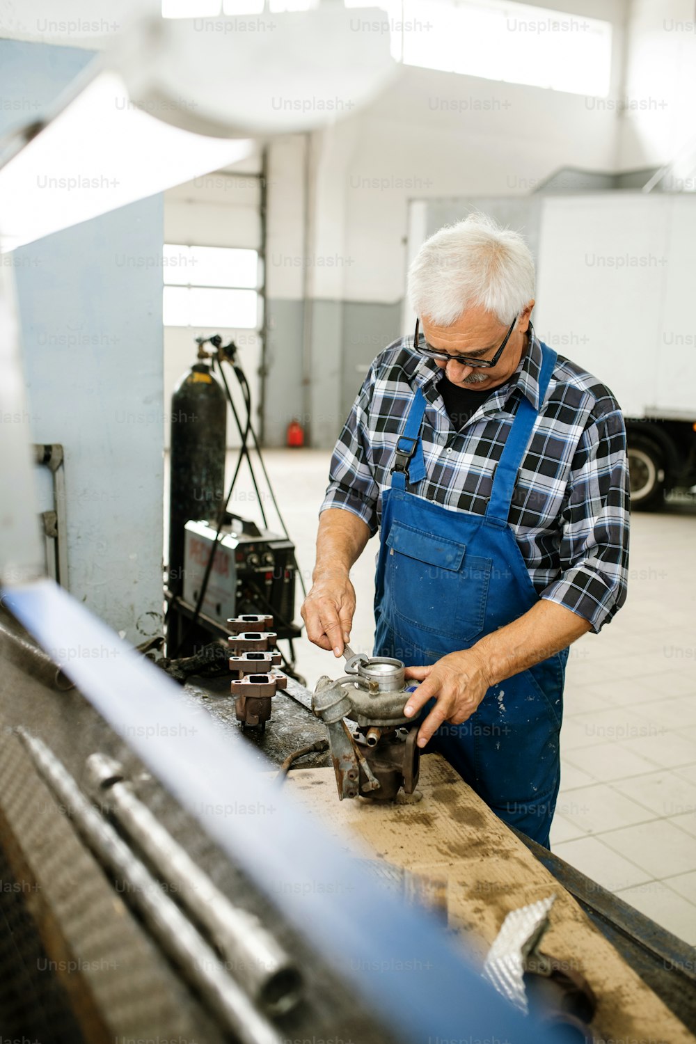 Leitender professioneller Techniker in Overalls und Hemd, der am Arbeitsplatz steht, während er einen Schraubenschlüssel verwendet, um Teile des Automotors zu reparieren