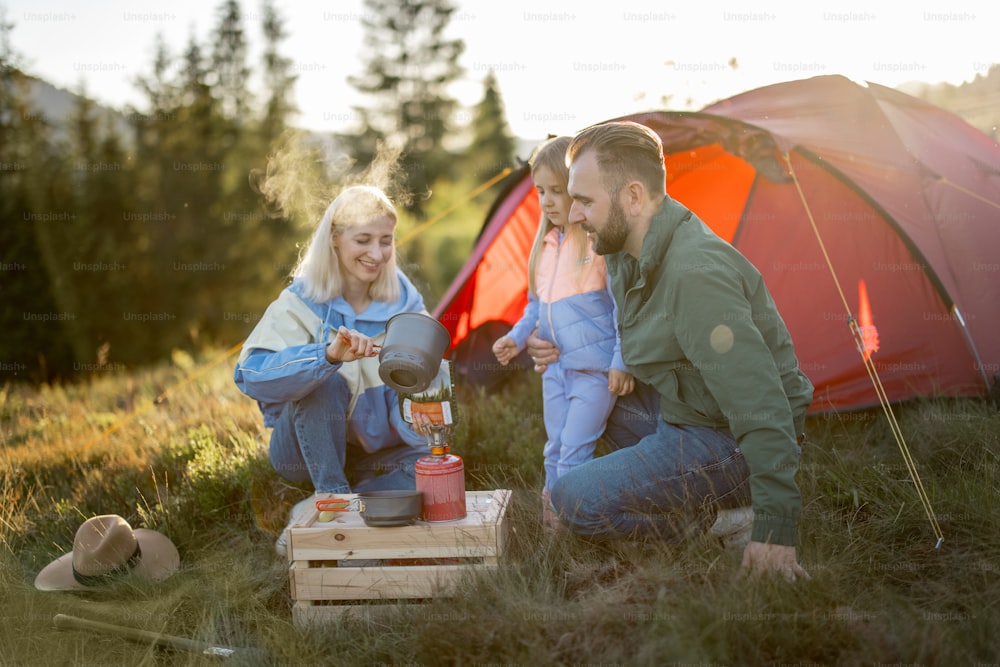 Couple with little girl have picnic at campsite while traveling with tent in the mountains. Young family happily spending vacations on nature