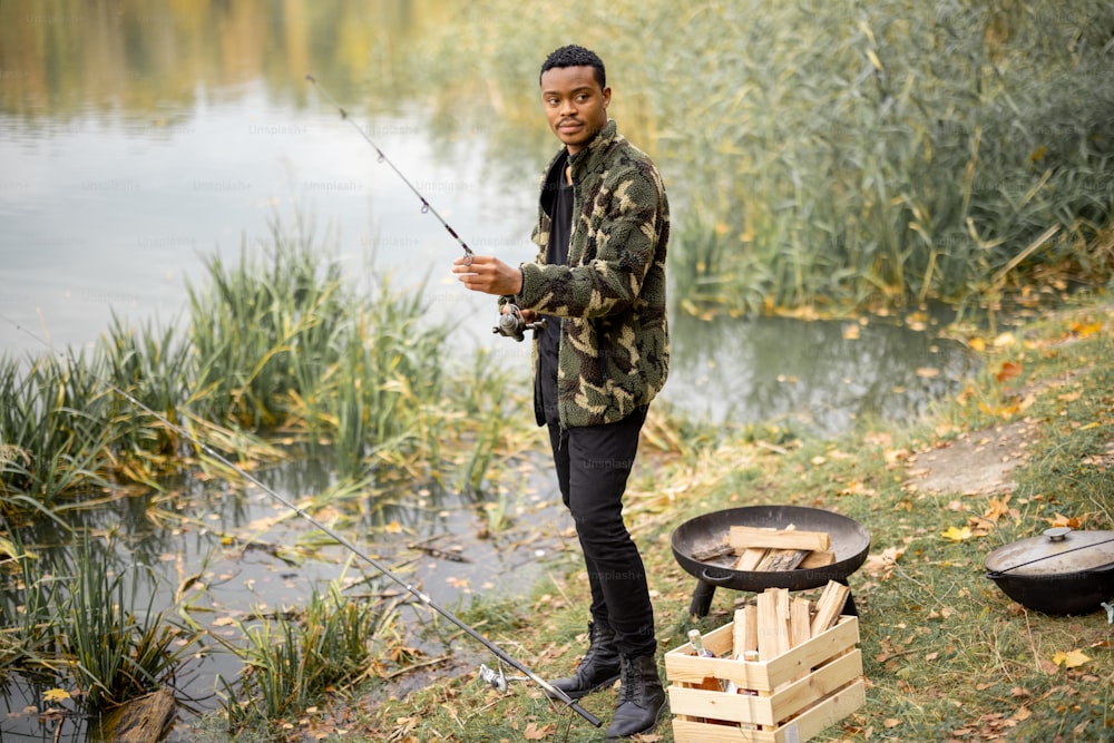 Porträt eines hispanischen Mannes, der in der Herbstsaison an der Fluss- oder Seeküste fischt. Konzept von Ruhe, Hobby und Wochenende in der Natur. Selektiver Fokus der männlichen Person in warmer Kleidung