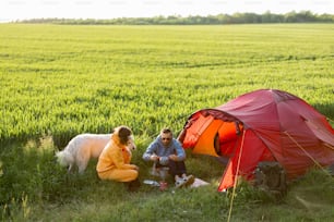 Young couple cooking food, spending summer time with a dog at campsite on the green field. Man and woman travel with tent on nature