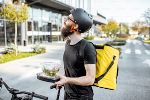 Coursier jeune et joyeux attendant un client avec des déjeuners à emporter près du bâtiment à l’extérieur, livrant de la nourriture de restaurant sur un vélo à l’aide d’un sac isotherme