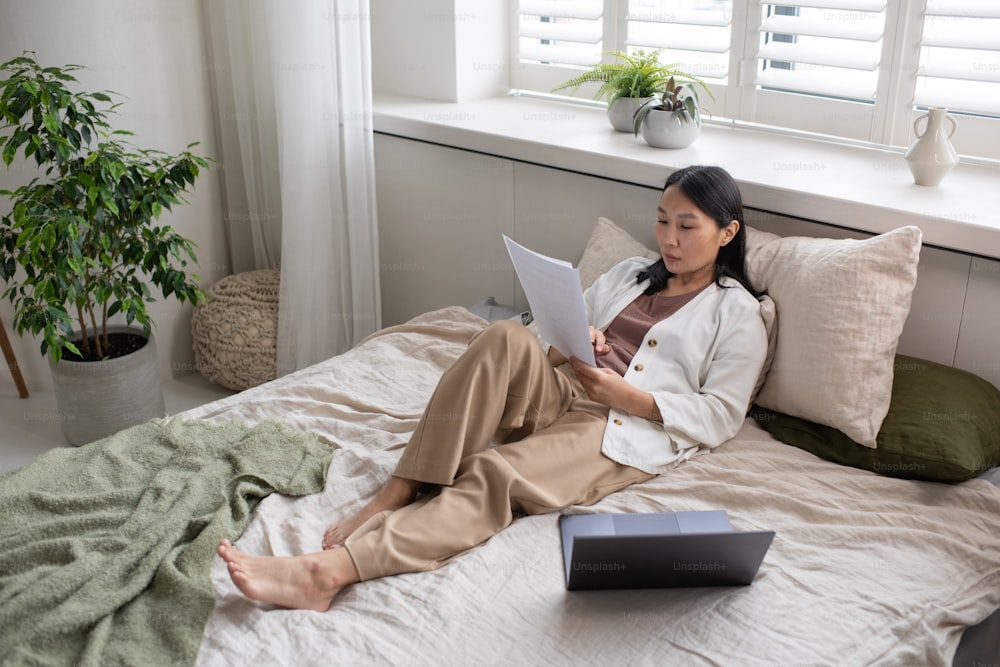 Young confident Asian female in casual clothes looking through financial papers while sitting on double bed against large window