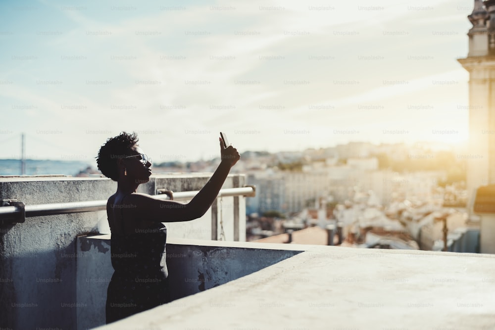 Uma silhueta de uma jovem negra de óculos tirando fotos em seu celular no telhado com uma paisagem urbana ensolarada ao seu redor; uma mulher africana está fazendo uma selfie através de um smartphone no telhado