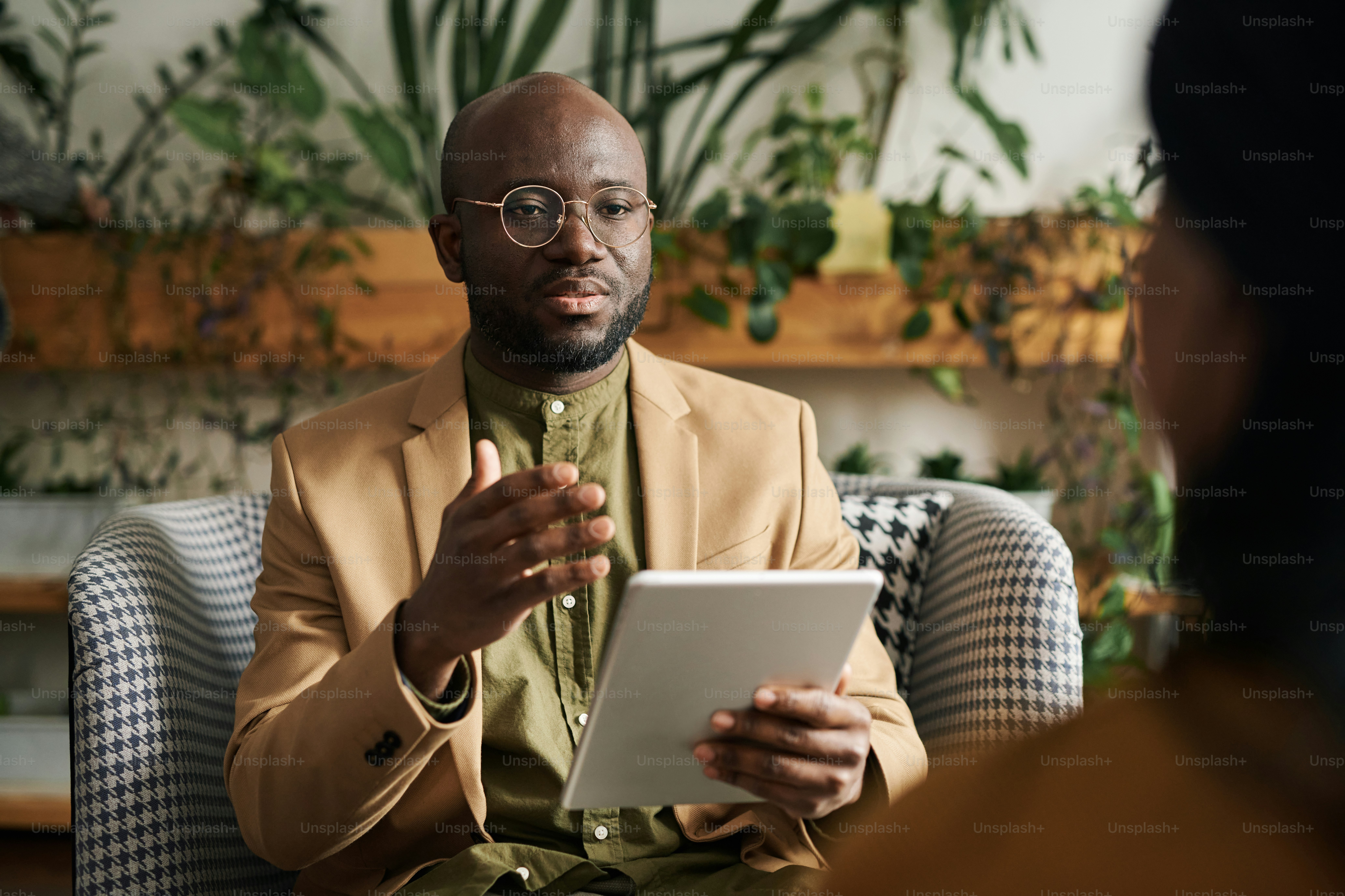 Selbstbewusster afroamerikanischer Berater mit Tablet, der eine Patientin berät, während sie während der psychologischen Sitzung vor ihr sitzt